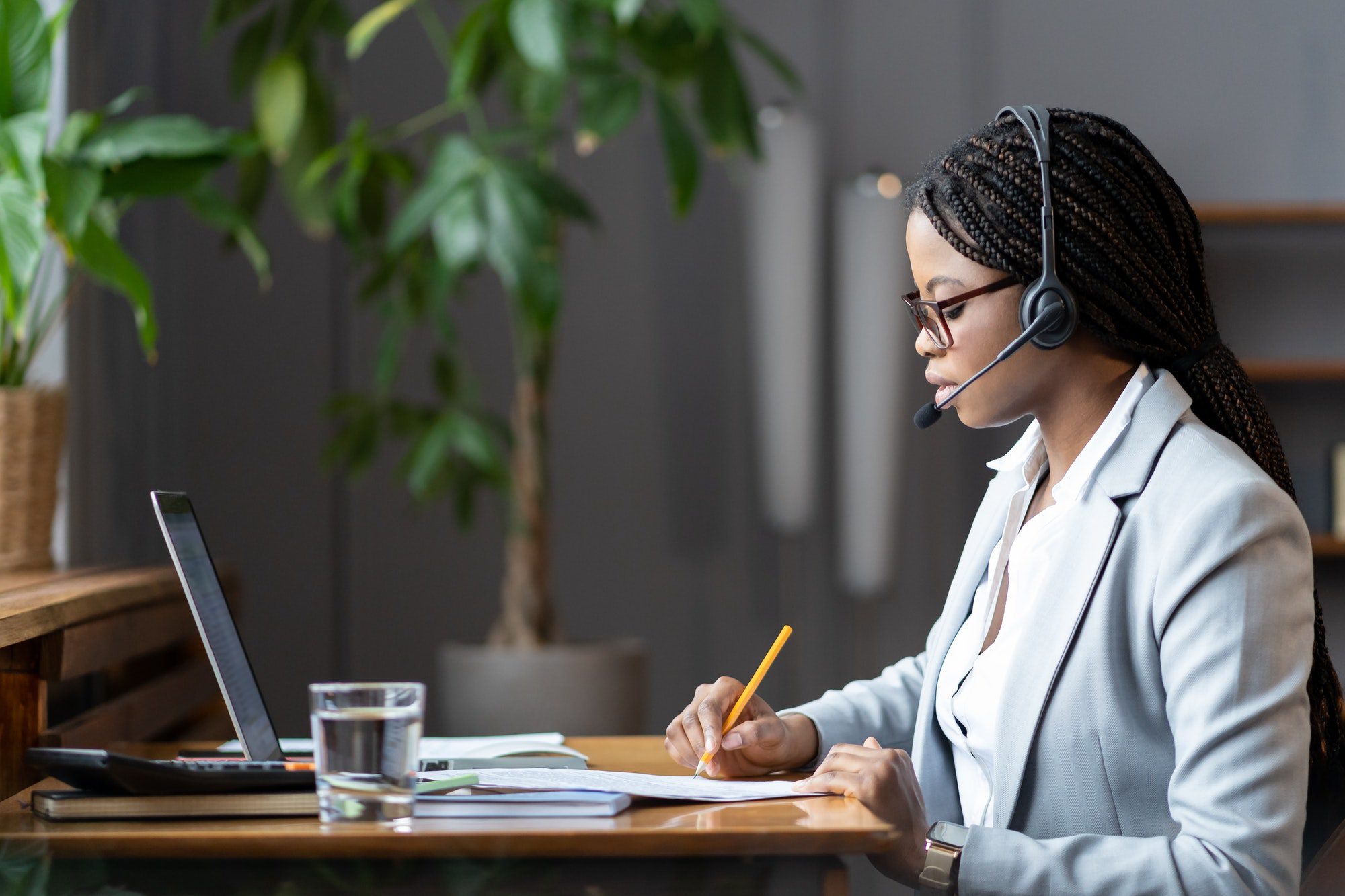 Focused african female home-based virtual assistant in headset making notes during online meeting